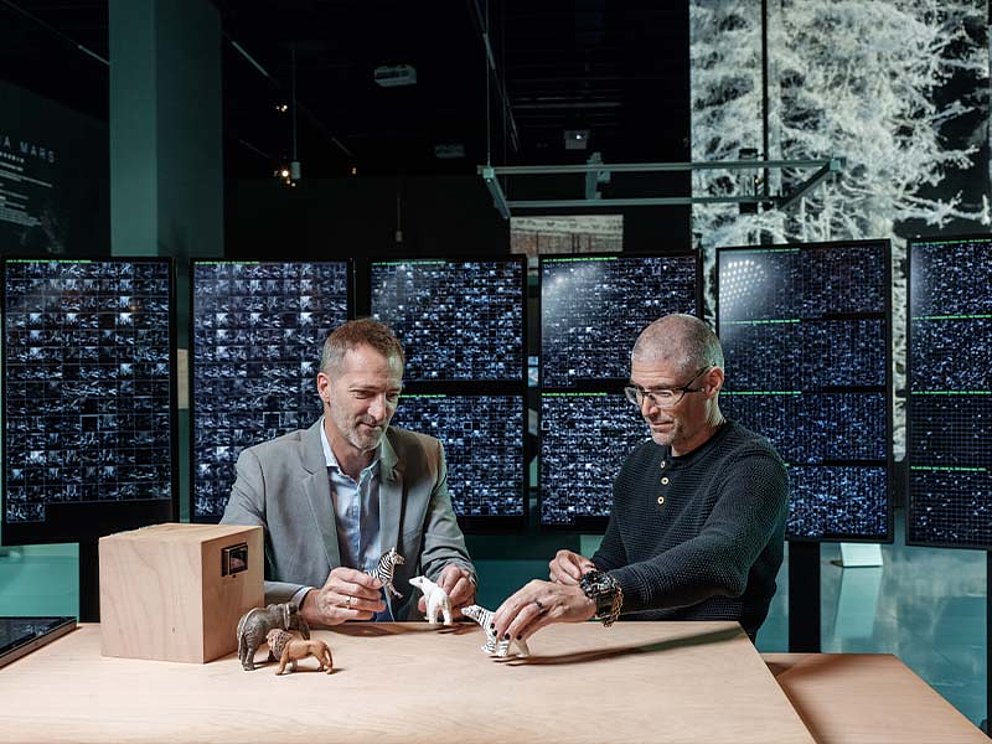 2 Männer sitzen am Tisch, spielen mit Tier-Figuren, im Hintergrund eine Monitor-Wand