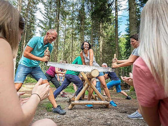 2 Personen beim Sägen eines Holzstammes, rundherum Menschen, die die beiden anfeuern.