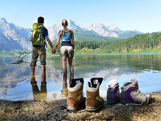 2 Wanderer bis zu den Knöcheln im Almsee, die Wanderschuhe am Seeufer, im Hintergrund Bergpanorama