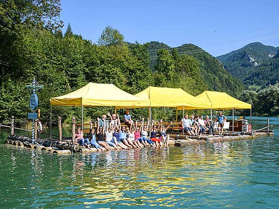 Eine Gruppe am Floß sitzend mit den Füßen im Wasser und winkend.
