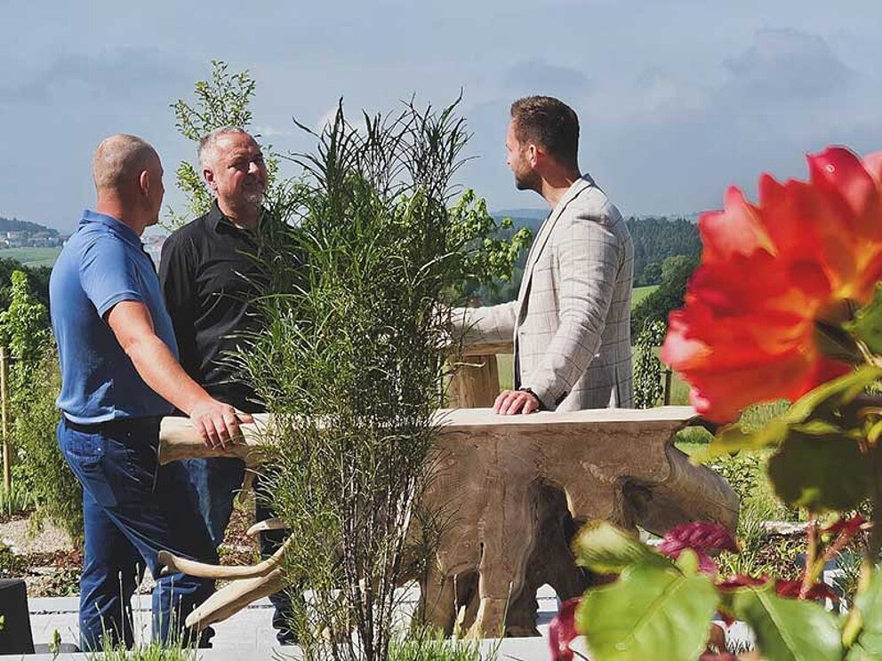 3 Männer stehen an einem Fass in einem blühenden Garten und verkosten Whisky