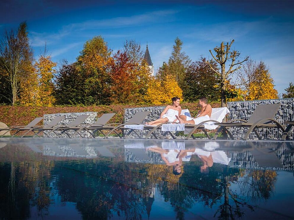 Ein Paar sitzt liegt auf Relaxliegen beim Außenpool, im Hintergrund herbstlich verfärbte Laubbäume.