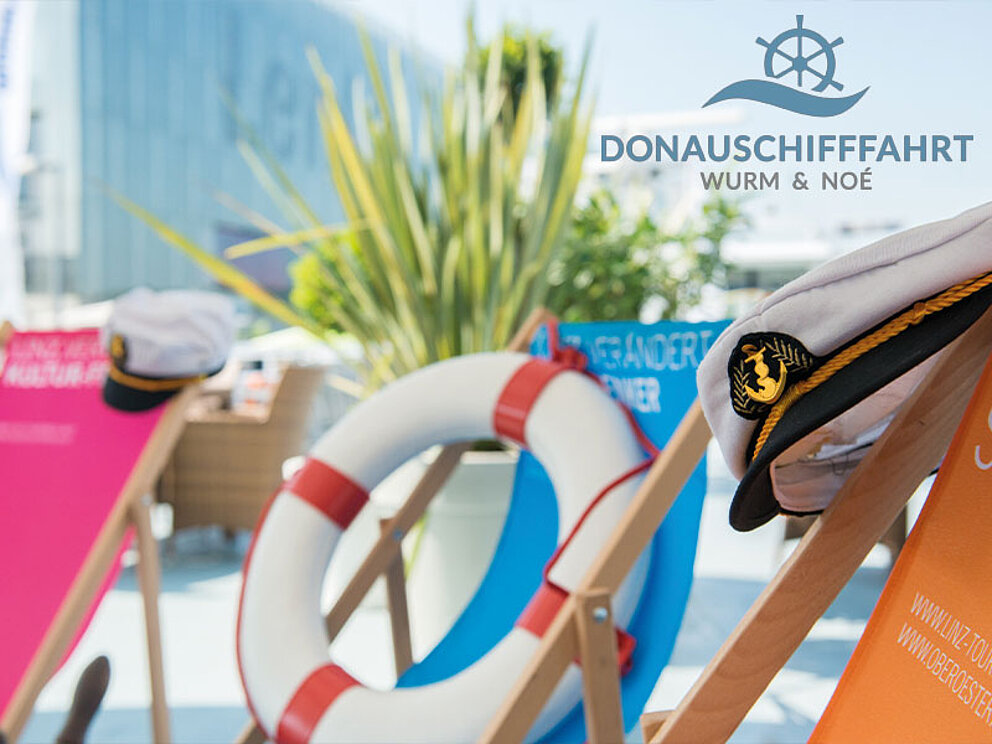 Eine Kapitänskappe hängt über einem Liegestuhl, in einem anderen liegt ein Rettungsring.