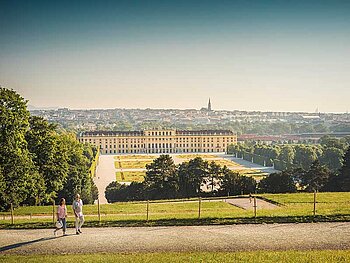 Paar spaziert im Schlosspark Schönbrunn. Im Hintergrund das Schloss und die Silhouette der Stadt.