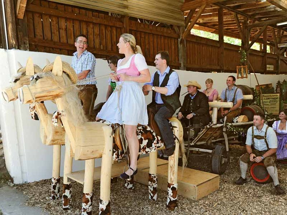 junge Leute in Tracht auf Holzpferden mit Kutsche im Stadel