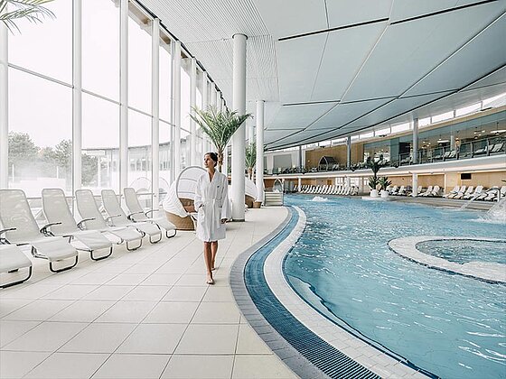 eine Dame im Bademantel geht entlang des Beckenrandes im Innenbereich der Therme