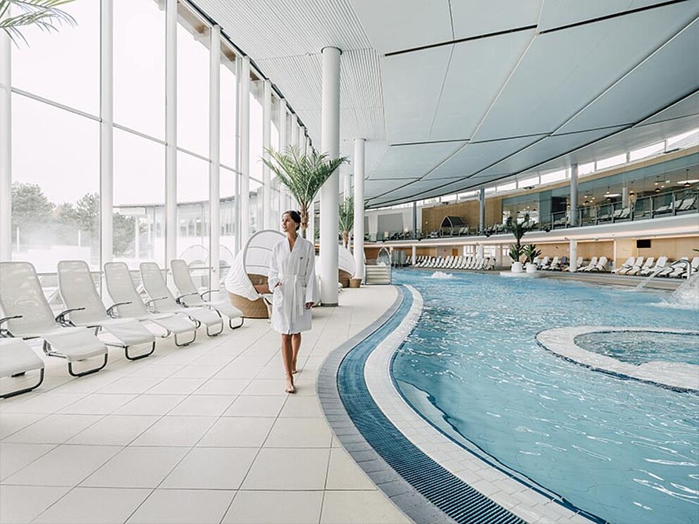 eine Dame im Bademantel geht entlang des Beckenrandes im Innenbereich der Therme