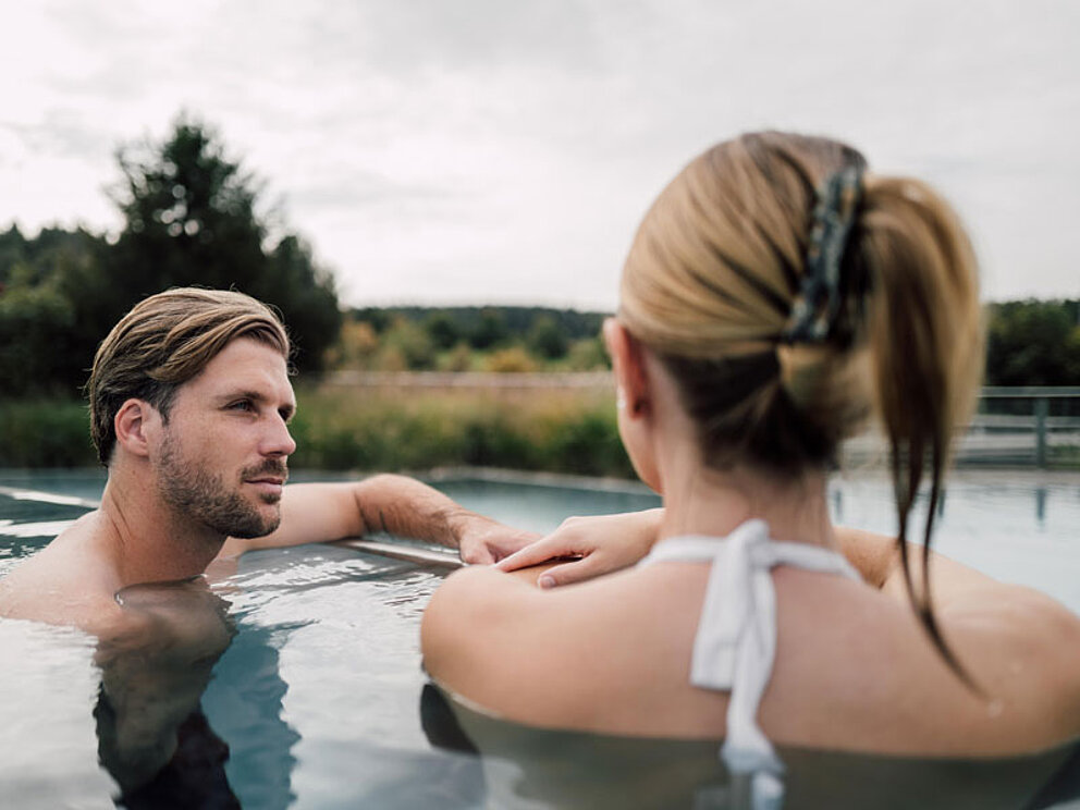 Mann und Frau liegen im Outdoor Thermalbecken und sehen sich an.