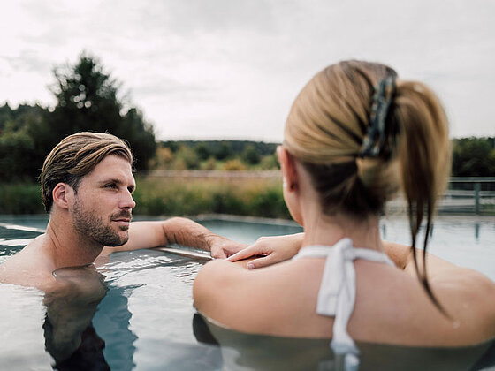 Mann und Frau liegen im Outdoor Thermalbecken und sehen sich an.