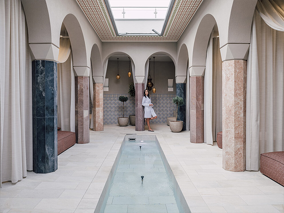 Eine Dame im Bademantel lehnt an einer Säule in einem überdachten Arkadenhof. In der Mitte ein rechteckiger Springbrunnen.