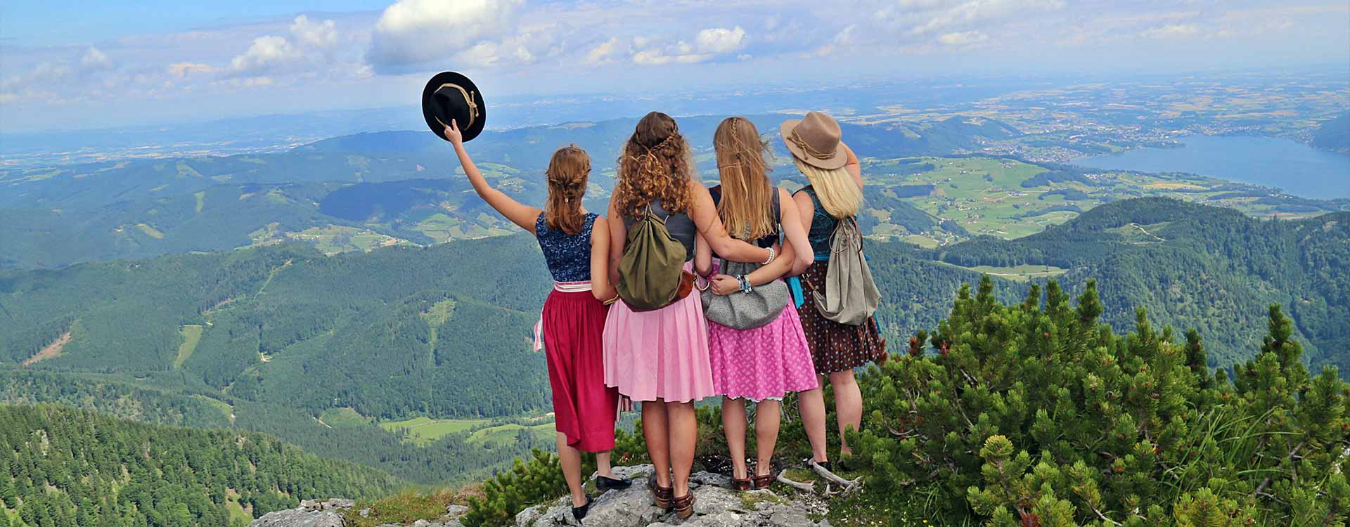 4 Damen im Dirndl von Hinten blicken vom Berg in die umliegende Landschaft und genießen das Panorama