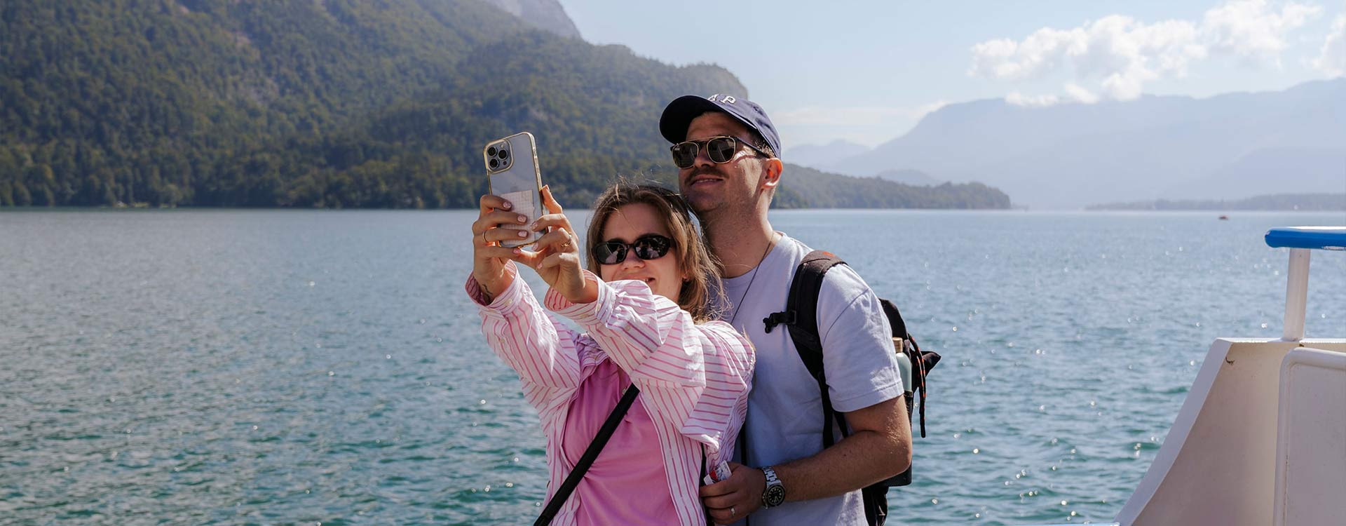 ein Paar macht ein Selfie von sich auf dem Wolfgangseeschiff, im Hintergrund der See und Berge